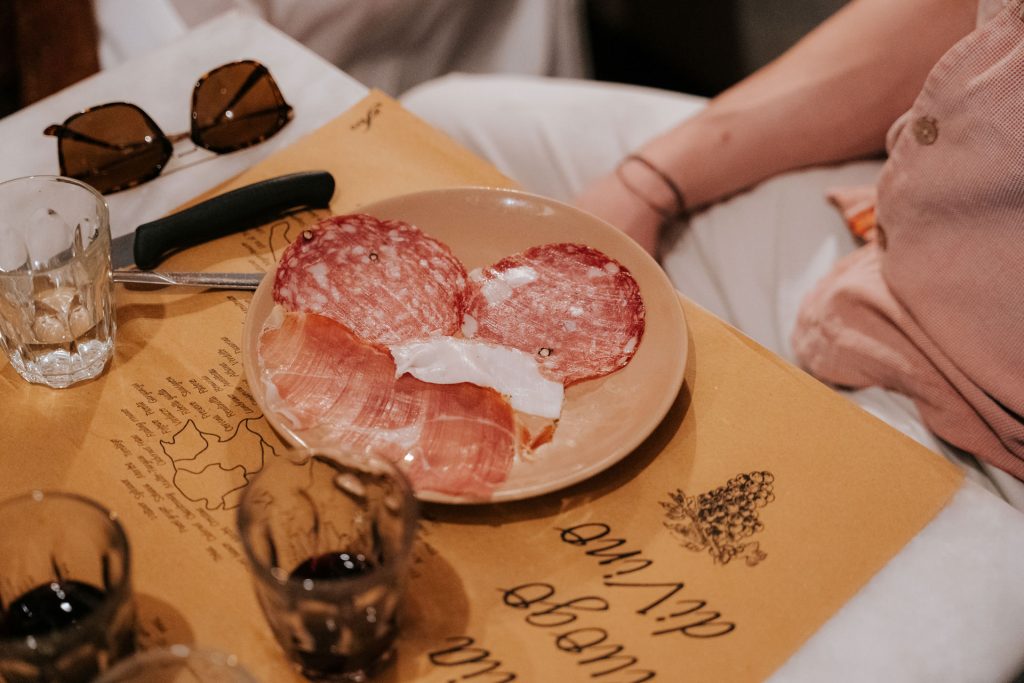 Several slices of cured meats and prosciutto rest on a plate atop brown paper, with empty glasses, cutlery, and sunglasses nearby. Cold cuts offer a traditional taste of Tuscany