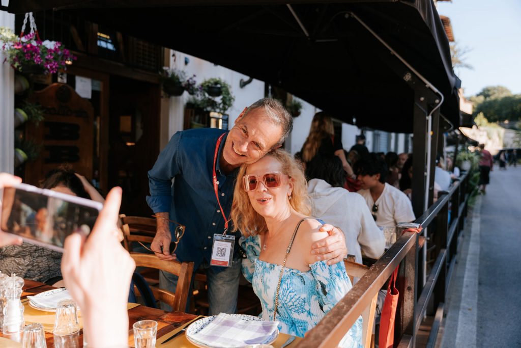 A man in a blue shirt and a woman in a blue dress with pink sunglasses smile together at an outdoor restaurant to enjoy a Taste of Tuscany menu