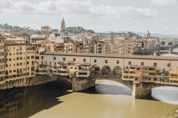 The Ponte Vecchio crosses the Arno River in Florence, with medieval shops and yellow buildings under a partly cloudy sky.
