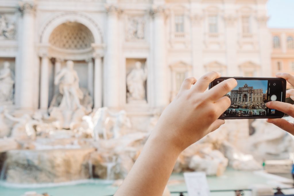 A person uses a smartphone to photograph Trevi Fountain in Rome, focusing on its ornate sculptures and cascading water. One of the tips for visiting Rome is to go underground and found the hidden parts of the city, including under the trevi fountain.