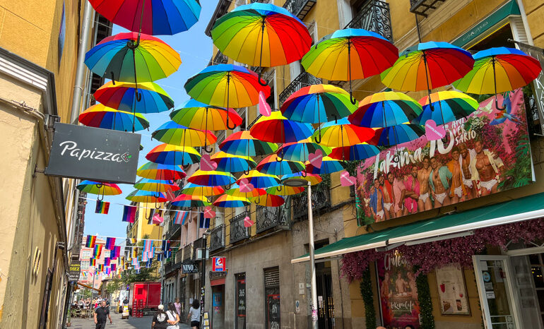 Colorful rainbow umbrellas and pride flags hang above a lively street in Chueca, the Madrid gay district, with LGBTQ+ venues and shops visible below. A welcoming barrio for solo travelers in Madrid