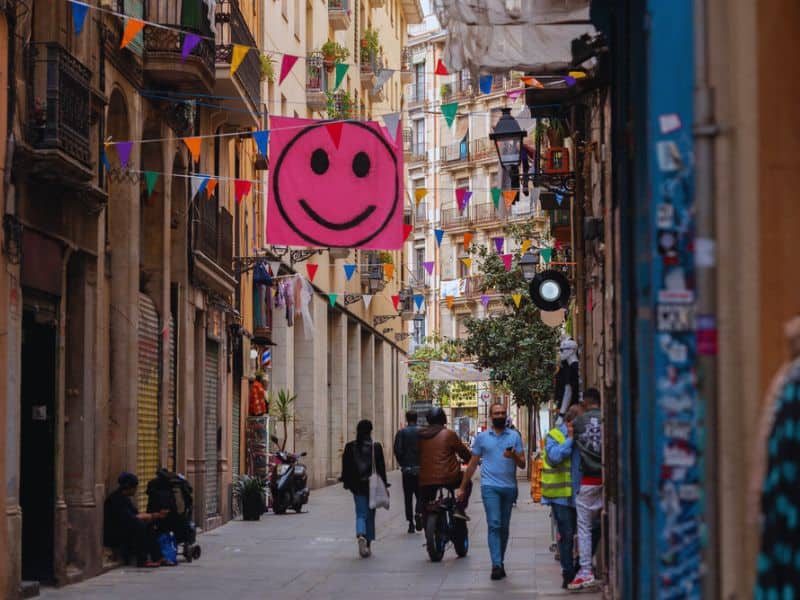 A lively street in the Barcelona gay neighborhood calle El Raval. The street features colorful banners, a large pink smiley sign, people walking and riding scooters.