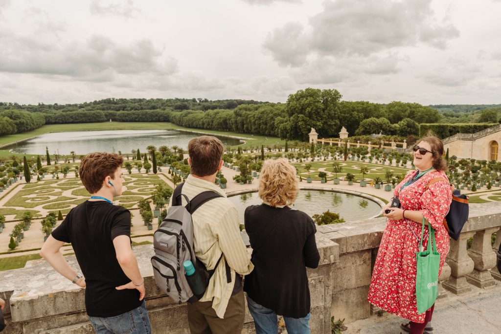 Four people stand on a stone terrace, listening to a woman in a red dress explain the grand formal gardens below.