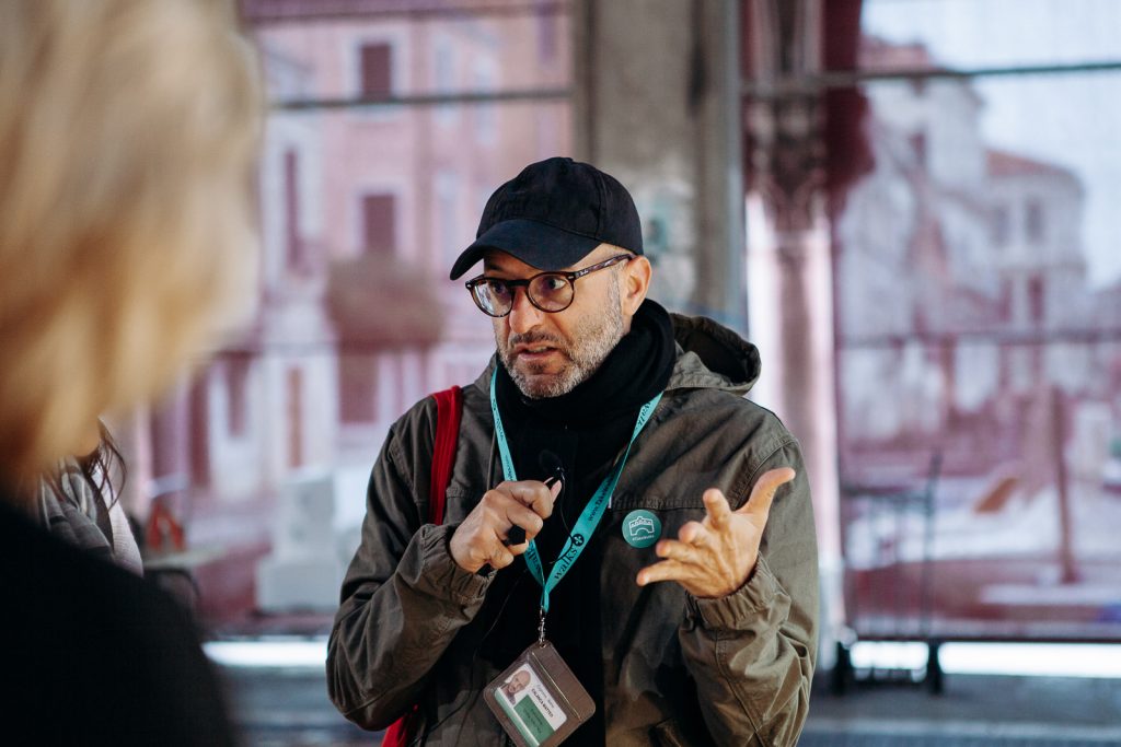 A man with glasses, black cap, and jacket speaks passionately into a microphone outdoors, with city buildings and bystanders behind him.