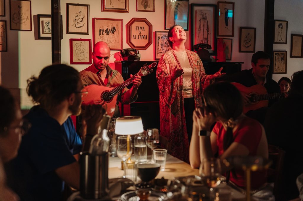 A singer performs Fado in Lisbon, with guitarists for an attentive group in a warmly lit restaurant decorated with framed art on the walls.