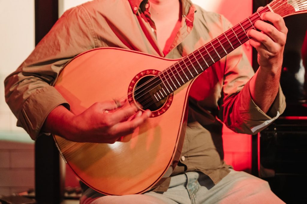 A person in a button-up shirt expertly plays a Portuguese guitar under warm red lights on stage, focused and expressive. Experiencing the music genre of Fado in Lisbon is a must for visitors who want to enjoy the cultural traditions of the city