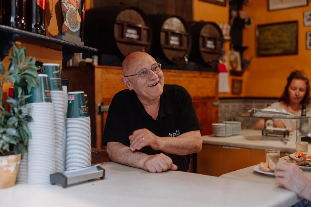 A smiling man in glasses chats behind a counter at one of the most famous tapas bars in Barcelona, . Cups and bottles are stacked nearby. A woman eats in the background.