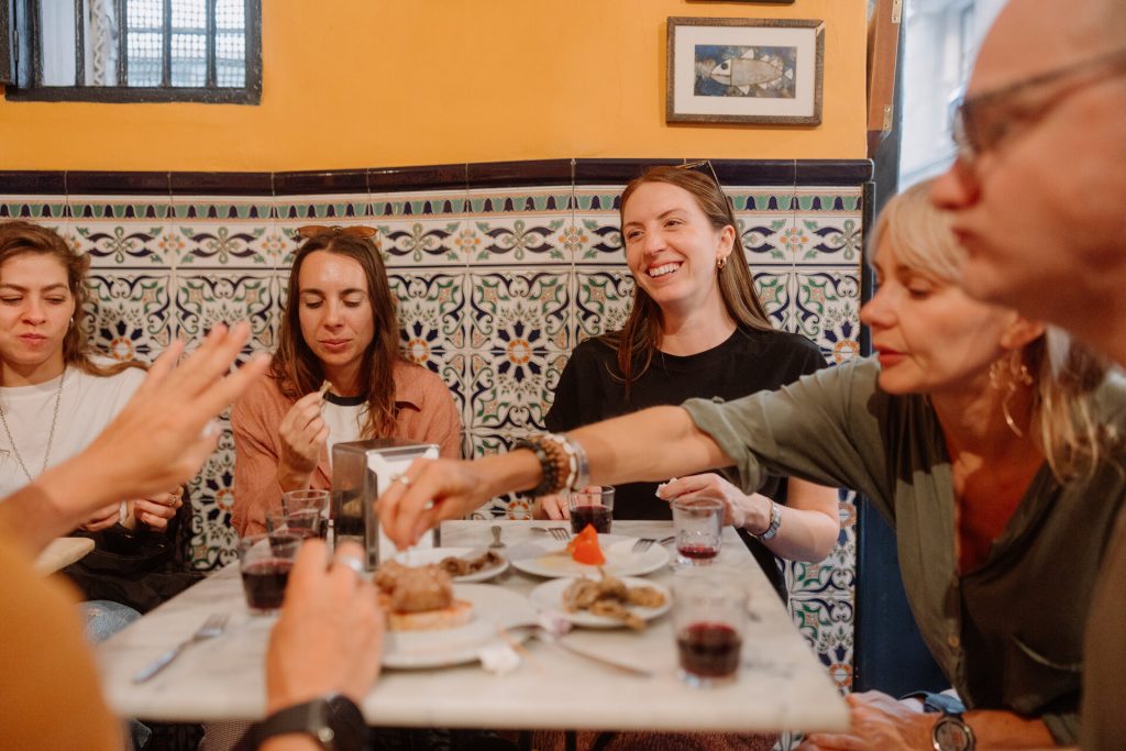 Several people share different plates at one of the local tapas bars in Barcelona, They laugh together at a table filled with food and drinks with vibrant tiled walls and framed artwork behind them.