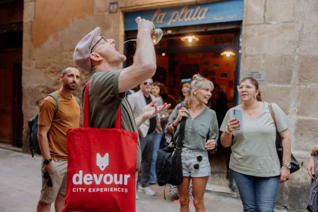 A man drinks wine from a glass pitcher outside one of the Tapas bars in Barcelona, Spain, watched by several people near stone buildings.