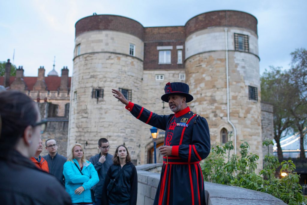 A Beefeater in traditional uniform leads attentive tourists outside the Tower of London as evening light casts shadows on ancient stone walls. Meeting a Beefeater is one of the top things that should be on your London Travel Checklist