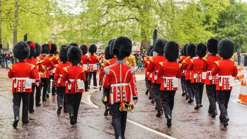 A photo of the Guard Change at Buckingham Palace. British guards in red uniforms and tall black hats march in formation on a wet road, watched by crowds and framed by trees.