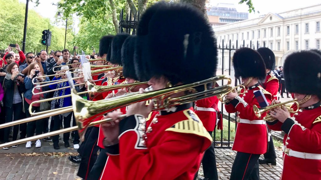 A photo of the Guard Change at Buckingham Palace. British guards in red uniforms and bearskin hats play trombones in a parade. Spectators watch behind a fence. Trees stand behind.
