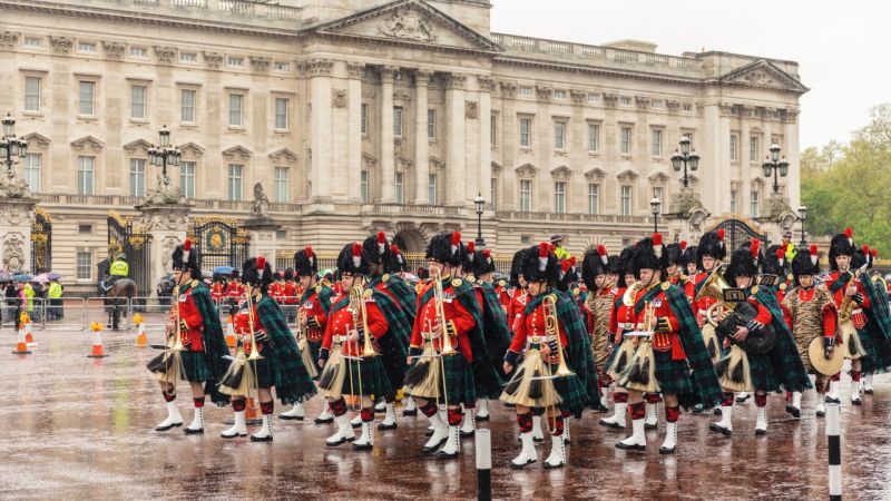 A Scottish marching band wearing kilts and feathered hats plays music outside Buckingham Palace during rainy weather. Witness this and other soldiers doing the Guard Change at Buckingham Palace.