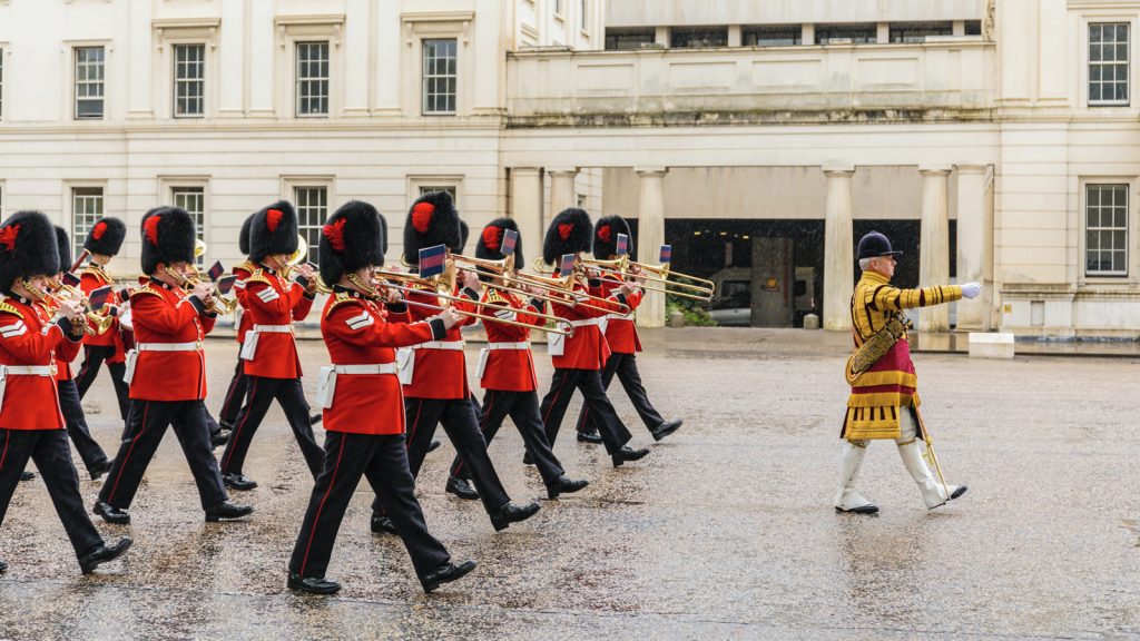 A photo of the Guard Change at Buckingham Palace. Soldiers in red uniforms and bearskin hats march in formation, playing brass instruments outside Buckingham Palace, London.