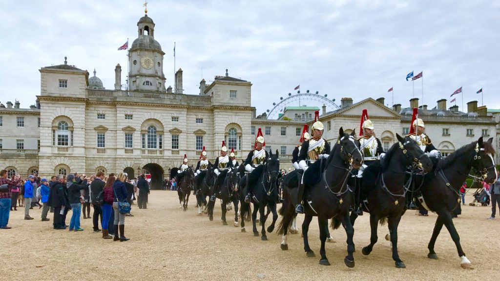 Mounted guards in ceremonial uniforms ride black horses past crowds near Horse Guards Parade, with the London Eye visible behind. Part of the traditional Guard Change ceremonies at Buckingham Palace.