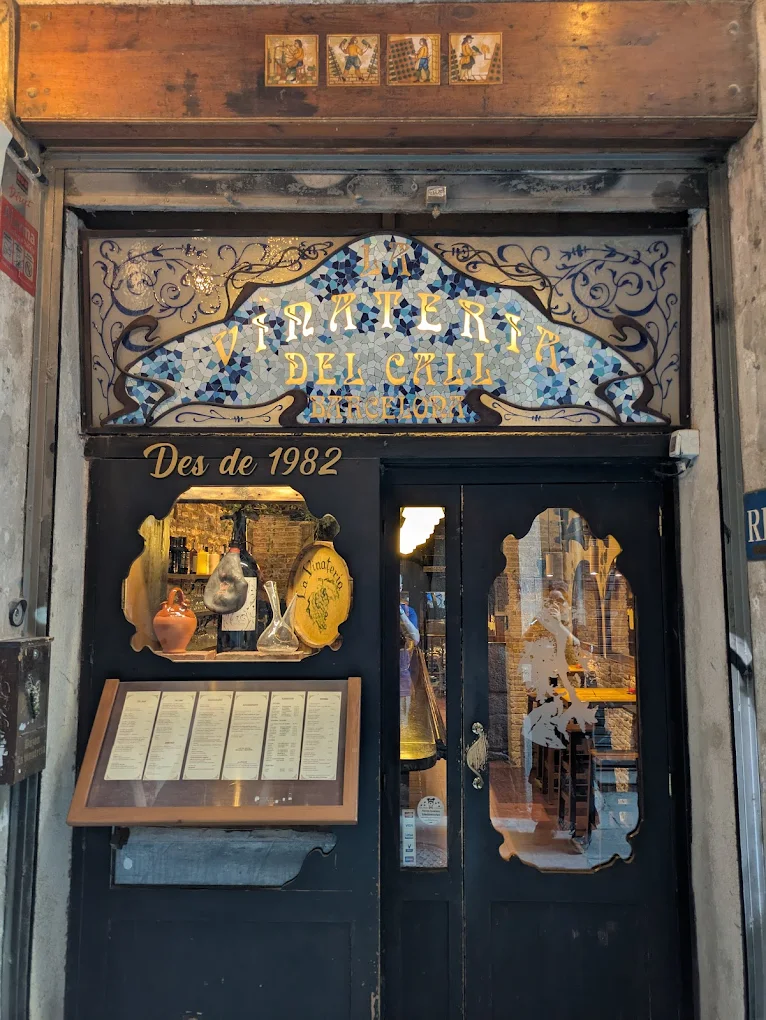 The entrance of Vini & Trine del Cali, a tapas bar in Barcelona, features ornate glass, gold-detailed doors, and menus by a sign from 1982.