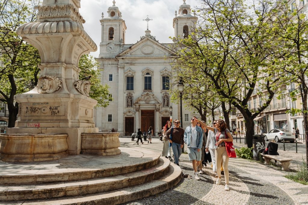 Several people stroll past a stone fountain in Lisbon’s sunny plaza, with leafy trees and a historic church behind them. Wandering the streets is the best way to see the sights on a solo trip to Portugal.