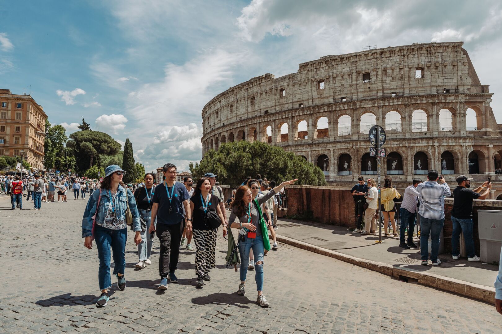Tourists walk and talk with a tour guide in front of the Colosseum in Rome on a sunny day, with other visitors and old buildings nearby.