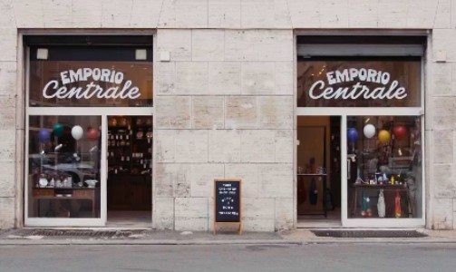 Two large gift-filled windows and Emporio Centrale signs appear on a stone building, with a black sandwich board between doors. The ideal place to buy gifts for travel lovers. 