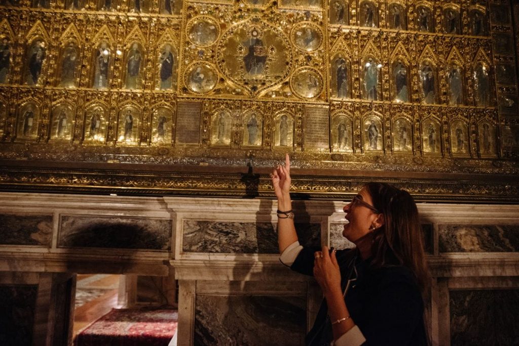 A woman with glasses gestures toward a gilded, intricate religious altarpiece with sculpted figures in a historical church interior.