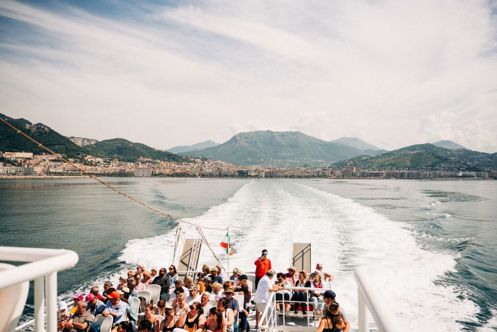 Several people stand on a boat’s deck heading away from a coastal city toward mountains, leaving a white wake behind.