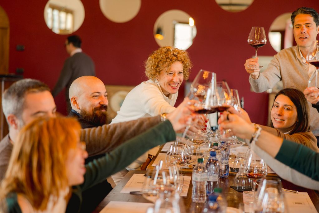 Friends around a table toast with wine, laughing together in a cozy room with a red wall, sharing travel stories.