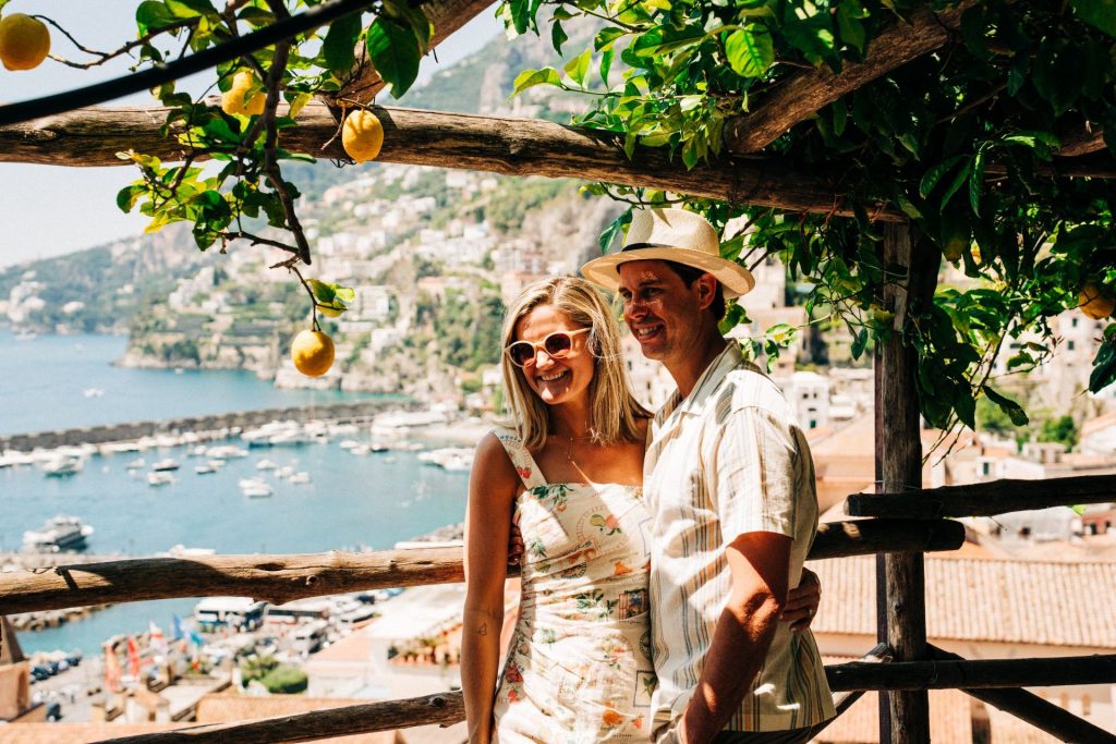 A smiling couple stands beneath a pergola with lemons, overlooking Positano’s marina, hillside buildings, and the bright blue sea.