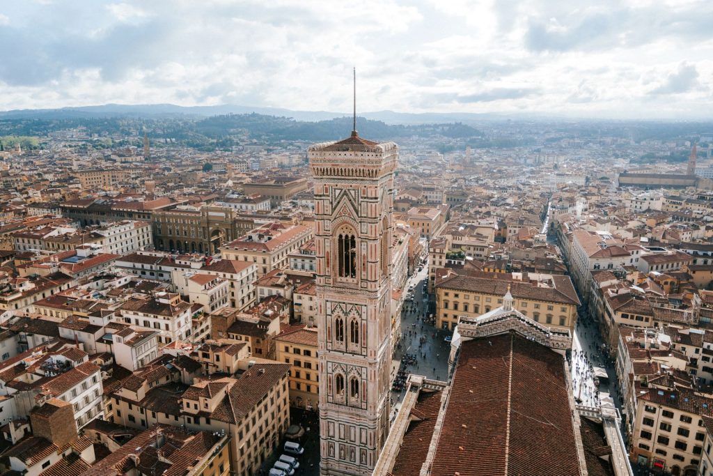 Giotto’s Bell Tower stands above Florence’s red rooftops, with distant hills and scattered clouds visible in the background.
