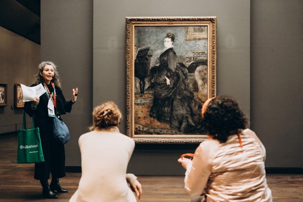 A museum guide explains Manet’s “Portrait of Berthe Morisot” to two seated visitors.