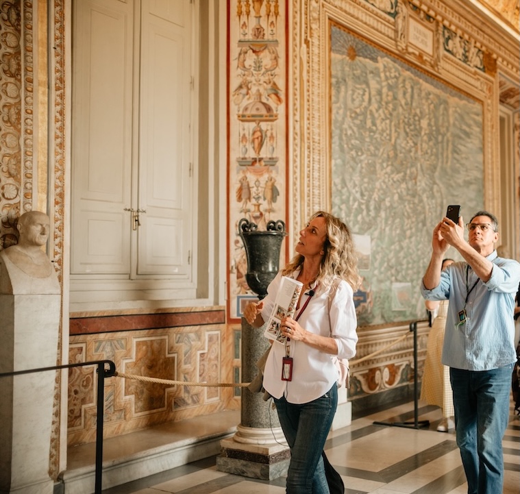 At the Vatican Museums, a woman holding brochures looks up as a man behind her photographs ornate statues and decorated walls.