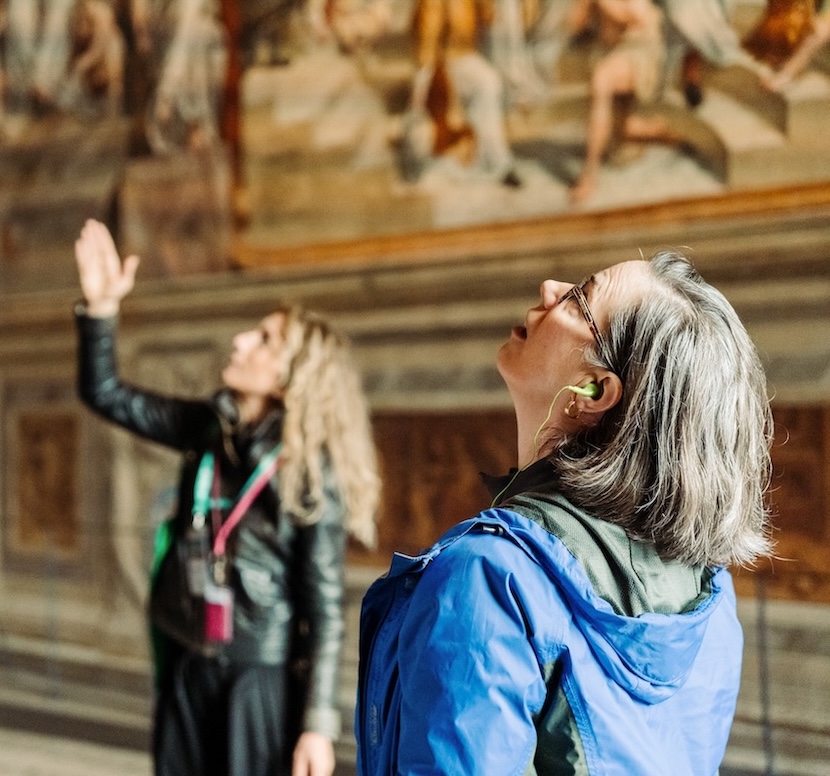 Two women observe a mural in the Vatican Museums; one points upward while the other listens to an audio guide.
