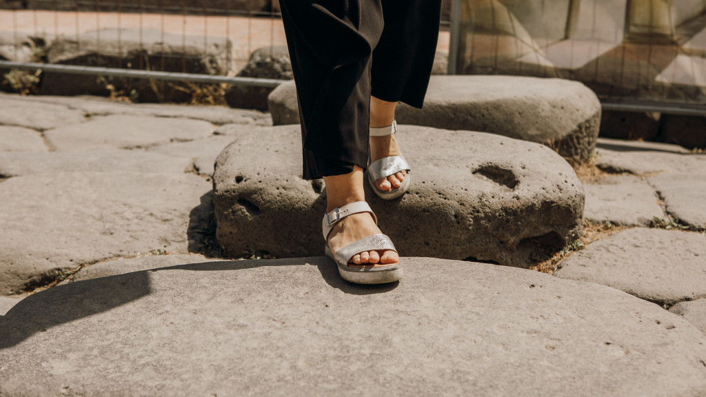 A person in white sandals and black pants walks across wide, ancient stone slabs outside, possibly at a historical site like Pompeii.