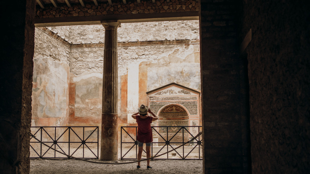 A visitor in a red shirt observes the ancient painted walls of Pompeii’s ruins from behind a metal fence under shelter.