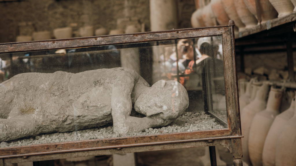 A plaster cast of a victim rests on its side in a glass case at Pompeii, surrounded by ancient pottery and artifacts.