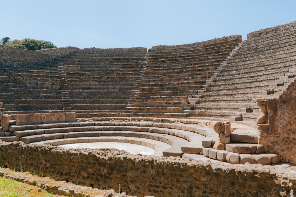 The ancient Pompeii amphitheater stands empty, its weathered stone seats under a clear blue sky, with scattered overgrown grass below.