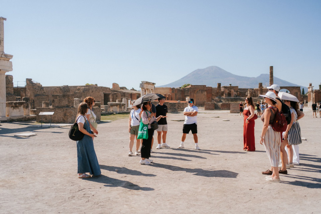 Tourists in summer clothes and hats listen to a Pompeii travel guide, with Mount Vesuvius rising in the background.