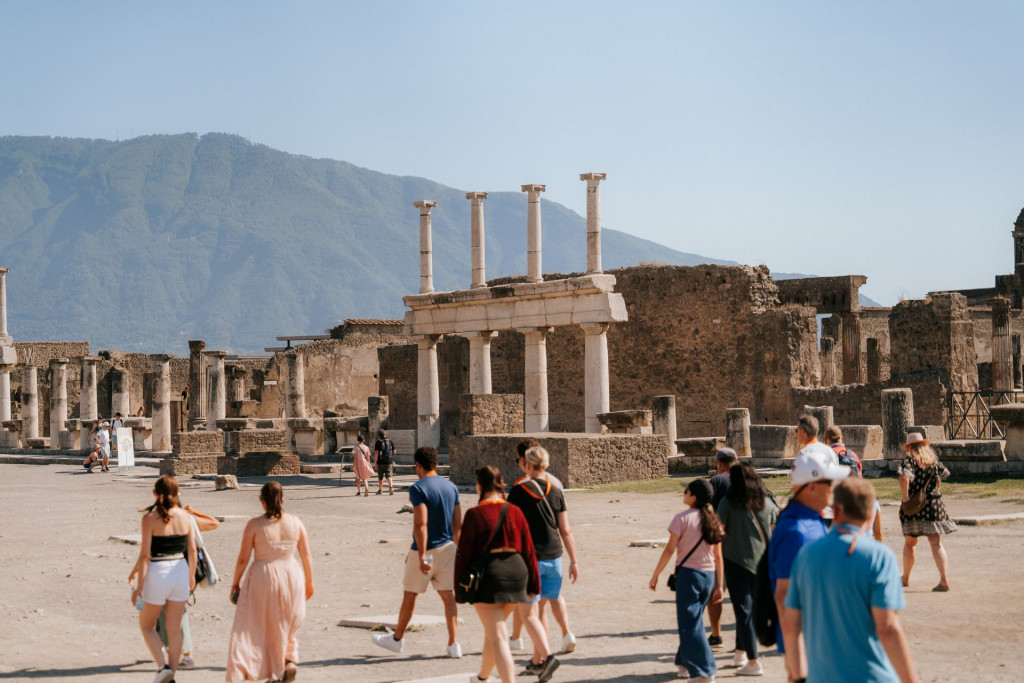 Tourists explore the ancient ruins with a Pompeii travel guide, passing tall stone columns while Mount Vesuvius rises in the clear background.