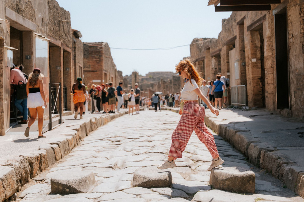 A woman with red hair walks across stepping stones on a street in Pompeii, surrounded by tourists and ancient ruins.