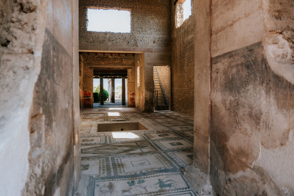 Sunlight illuminates a Pompeii stone interior with mosaic tile floor, sunken area, open windows, and a metal staircase upstairs.