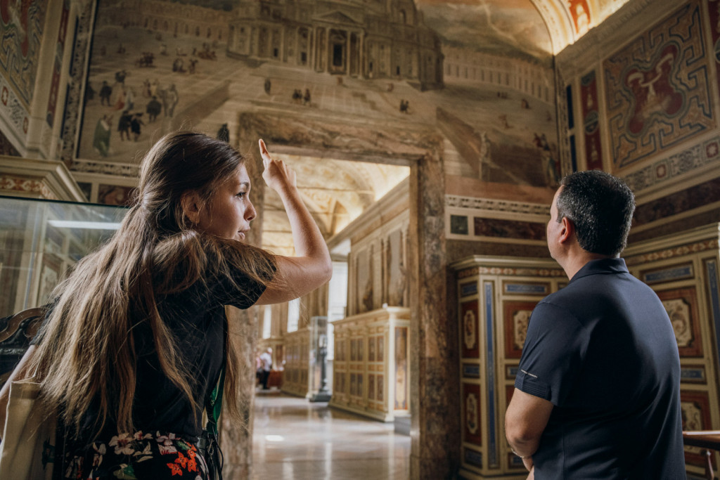 A woman gestures as she talks to a man inside the ornate Vatican Museums hall, with intricate frescoes and arched, patterned walls.