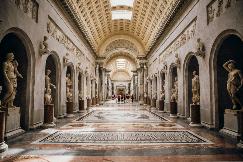 Visitors walk down a marble hallway in the Vatican Museums, admiring classical statues in alcoves beneath an ornate arched ceiling.