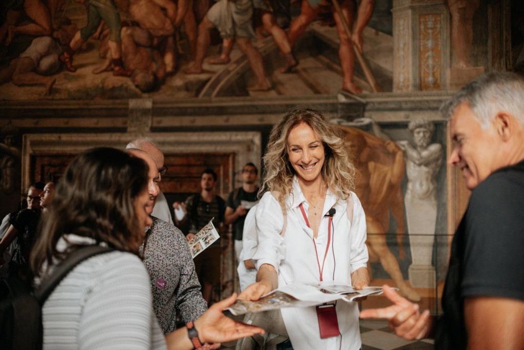 A smiling tour guide distributes brochures to visitors under Raphael’s School of Athens fresco in the Vatican Museums.